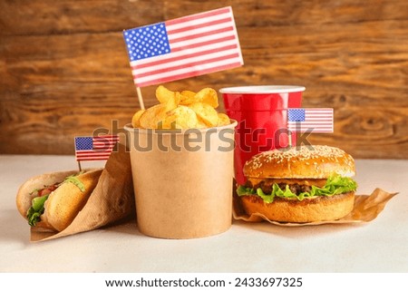Tasty burger, potato chips, hot dog and cup of drink on light table against wooden background. Memorial Day celebration