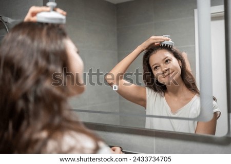 Young woman with dark curly hair doing self hair scalp massage with scalp massager or hair brush for hair growth stimulating at home bathroom. Reflected view of the mirror. High quality photo