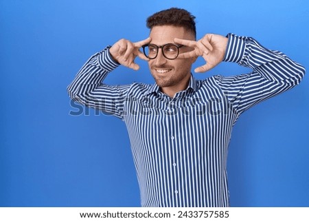 Hispanic man with beard wearing glasses doing peace symbol with fingers over face, smiling cheerful showing victory 