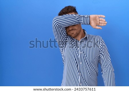 Hispanic man with beard wearing glasses covering eyes with arm, looking serious and sad. sightless, hiding and rejection concept  Royalty-Free Stock Photo #2433757611