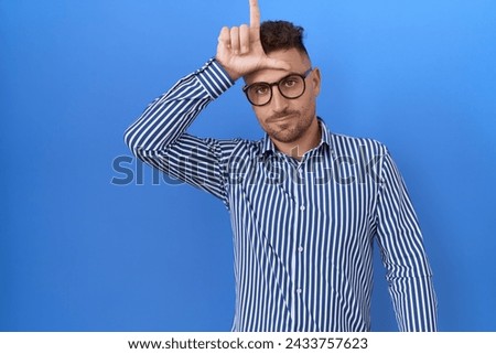 Hispanic man with beard wearing glasses making fun of people with fingers on forehead doing loser gesture mocking and insulting. 