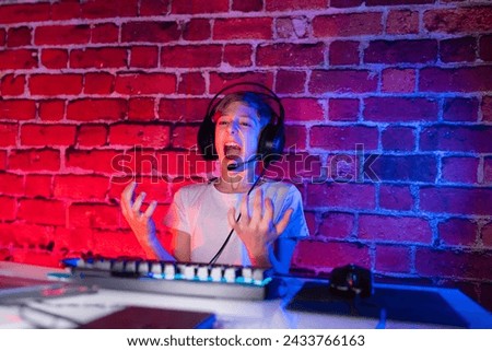 A teenager swings his arms around during video games. A gamer boy plays computer games against a neon lit brick wall.