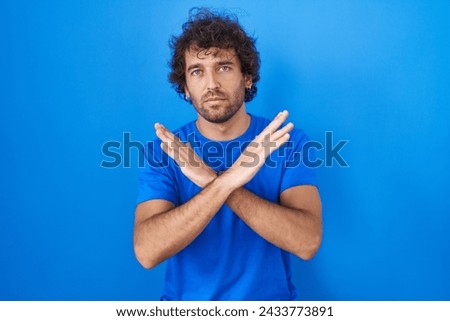 Hispanic young man standing over blue background rejection expression crossing arms doing negative sign, angry face  Royalty-Free Stock Photo #2433773891