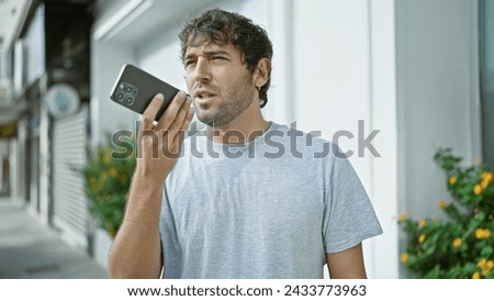 Relaxed yet serious, a young, blond, handsome guy with a beard is having an engaging, high-tech talk on his smartphone, sending a voice message outdoors on a bustling city street. Royalty-Free Stock Photo #2433773963