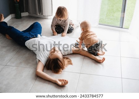 Young tired mother lying on floor relaxing or sleeping while her children drawing nearby. Exhausted woman with small kids at home. Bored female person with toddlers. Postpartum motherhood burnout.