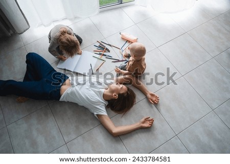 Young tired mother lying on floor relaxing or sleeping while her children drawing nearby. Exhausted woman with small kids at home. Bored female person with toddlers. Postpartum motherhood burnout. Royalty-Free Stock Photo #2433784581