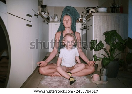 Modern yogini mom meditates while toddler sits on her lap at kitchen . Morning routine of working mother. Calm woman struggling with stress meditating in lotus pose on floor at home.