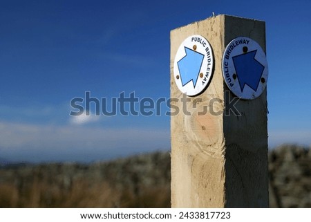 A wooden post has a pale blue arrows that points out the direction of a public bridelway. A polariser was used to enhance the blue colour of the sky. Royalty-Free Stock Photo #2433817723