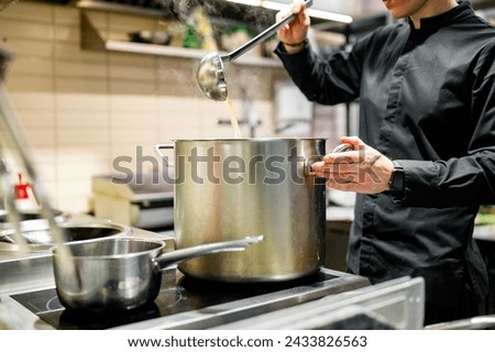 A chef in black attire stirs a large pot on a stove in a professional kitchen. Steam rises from the pot, indicating hot food. Other pots and pans are visible on the stove Royalty-Free Stock Photo #2433826563