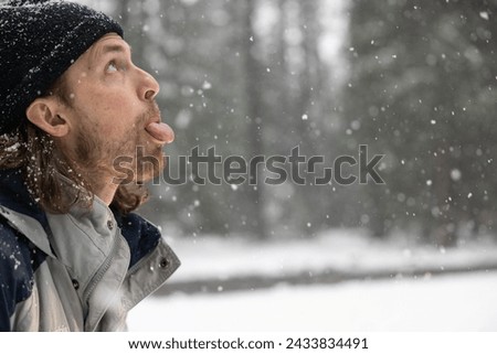 A man with a black hat and beard is sticking his tongue out in the snow. Concept of humor and playfulness, as the man is making a silly face in the midst of a snowy landscape Royalty-Free Stock Photo #2433834491