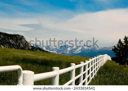 White rail fence with green grass and mountains in background.  Colorado high country scene of rural area.  Blue sky and white clouds. White fence with hills and mountains in the background.   Royalty-Free Stock Photo #2433834541