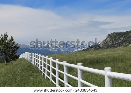 White rail fence with green grass and mountains in background.  Colorado high country scene of rural area.  Blue sky and white clouds. White fence with hills and mountains in the background.   Royalty-Free Stock Photo #2433834543