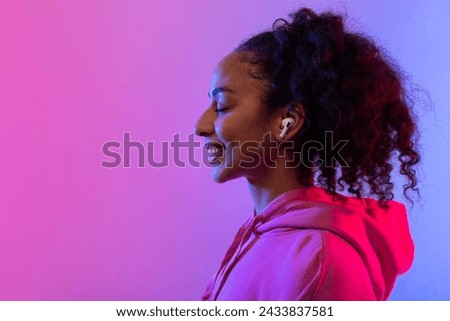 Smiling black lady with curly hair wearing earbuds, eyes closed in pleasure, against radiant neon pink and blue background, feeling the music, side view, free space Royalty-Free Stock Photo #2433837581