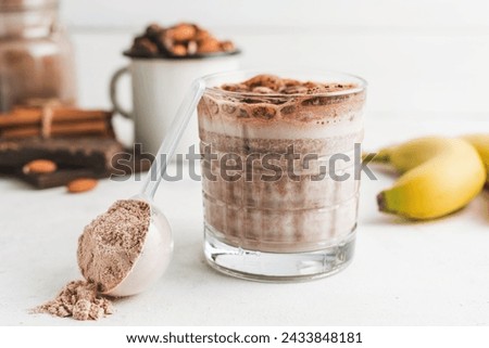 Glass with chocolate protein drink, milkshake smoothie on white table with bananas, protein powder in measuring spoon, protein bar, almond nuts and cinnamon sticks.