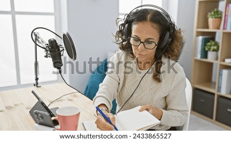 Mature hispanic woman recording podcast in home studio with microphone and notebook
