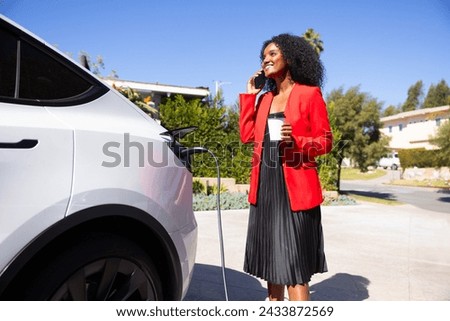 A professional african american woman charges her electrical car in the station standin on a very hot and sunny day while holding her cup of coffee and calling someone Royalty-Free Stock Photo #2433872569
