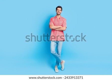 Full size photo of cool handsome man dressed striped shirt denim pants holding arms legs crossed isolated on blue color background