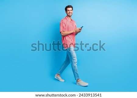 Full size photo of busy pleasant man dressed striped shirt denim pants hold smartphone go to empty space isolated on blue color background