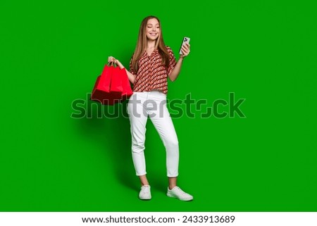 Full size photo of cute teenager wear print blouse hold shopping bags read notification on smartphone isolated on green color background