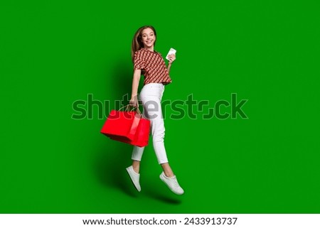 Full length photo of good mood girl dressed print shirt hold smartphone new outfit in bags jumping isolated on green color background
