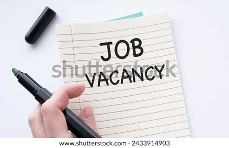 Close-up image of a person holding a black marker pen in hand over an open notebook with JOB VACANCY text written on a white page. Employment and recruitment concept image.