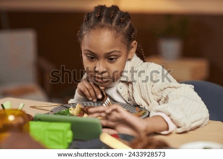 Front view of little African American girl using smartphone and watching videos while eating at dinner table