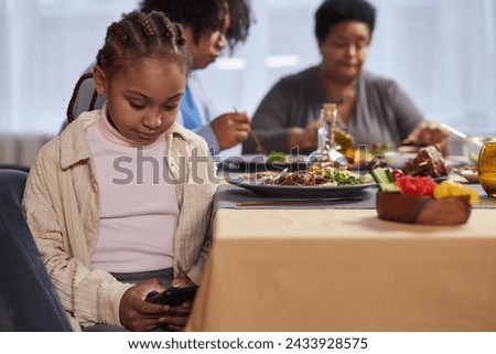 Portrait of young Black girl using smartphone under table at dinner and typing text messages copy space 