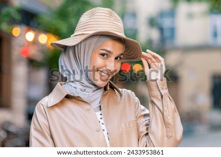 Portrait of attractive muslim woman wearing hijab and touching straw hat with playful facial expression. Delighted lady combining headwear and feeling ready for vacation season in stylish outfit. Royalty-Free Stock Photo #2433953861