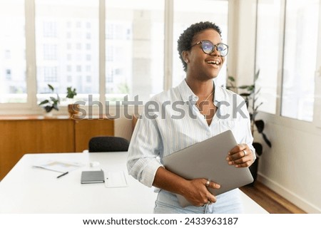 Successful cheerful inspired young african-american female entrepreneur, small business owner, female office employee, black businesswoman standing with laptop and laughing, real woman with teeth gap