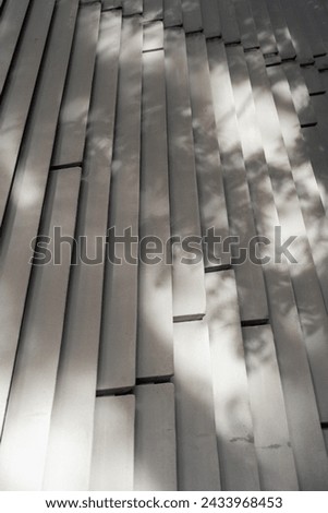 Surface of outdoors wall covered with vertically aligned and evenly spaced gray slats. Natural sunlight illuminating it and casting shadows between and on material