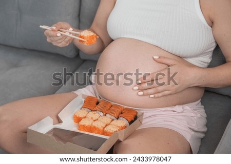 A pregnant woman sits on the sofa and eats rolls from a box. Food delivery. Close up of belly.  Royalty-Free Stock Photo #2433978047