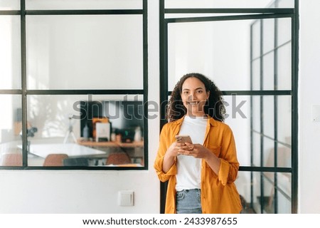 Chatting online. Happy brazilian or hispanic curly haired young woman in casual wear, stands at a modern office space, holding her mobile phone, chatting online, looks at camera, smiling friendly