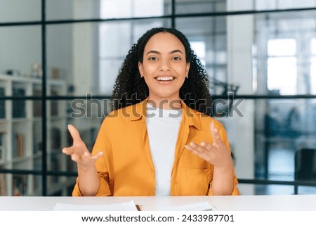 Confident pretty hispanic or brazilian curly haired young woman, talking via video call, listens or conducts an online lesson, coaching session, lecture, brainstorm, gesturing with hands, smiles