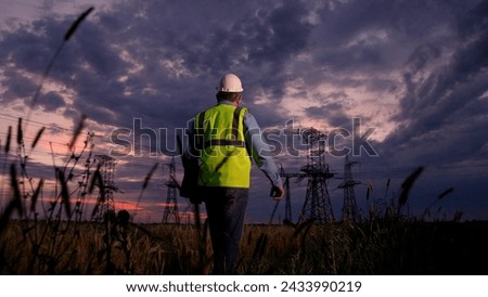 Power engineer in protective helmet on construction of power line using laptop, inspection, maintenance, power system. High voltage electrical lines at sunset. Construction engineer working outdoors Royalty-Free Stock Photo #2433990219