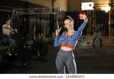 Woman female gym visitor takes pictures of herself on phone, takes selfie in gym. Slender and active girl in tight sports clothes looks at camera during photo shooting on smartphone camera