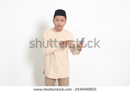 Portrait of surprised Asian muslim man in koko shirt with skullcap holding mobile phone, showing shocked face expression. Advertising and social media concept. Isolated image on white background