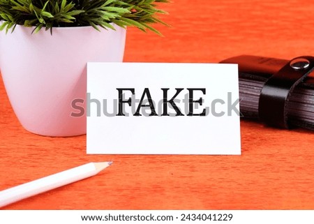 FAKE word written on a business card next to a business card holder, a green flower and a pencil on an orange background
