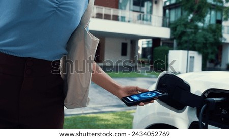 Modern woman recharge her EV car from home charging station by EV charger, checking battery status from EV smartphone app. Future lifestyle of sustainable clean energy utilization. Peruse