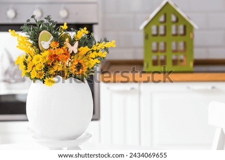 A spring bouquet in an egg, Easter bunnies and eggs with a golden pattern on the table. In the background is a white Scandinavian-style kitchen. A beautiful greeting card. Easter decor in the house.