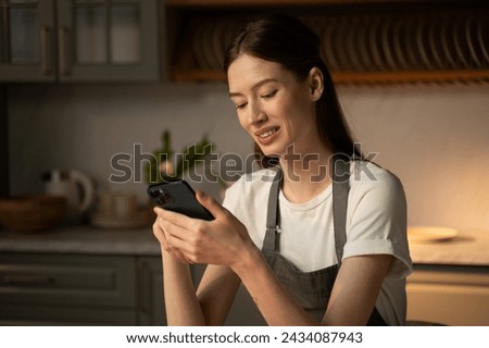 A young woman in a casual apron is sitting in a well-lit modern kitchen, staring intently at her smartphone. The soft glow of sunlight complements the warm atmosphere of the room, watching content on