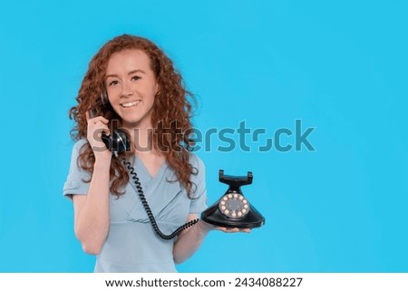 a smiling redhead woman in blue dress  speaking by vintage phone  and sitting on stool on a blue background with copy space. Happy people