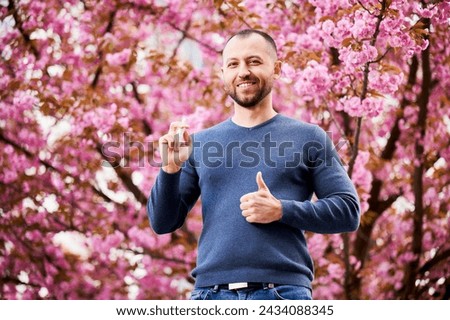 Man allergic using medical nasal drops, suffering from seasonal allergy at spring in blossoming garden. Portrait of smiling man showing thumbs up near blooming tree outdoors. Spring allergy concept. Royalty-Free Stock Photo #2434088345