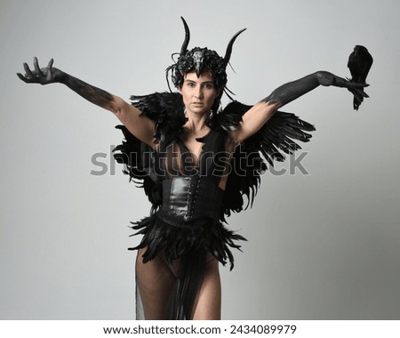 Close up portrait of female model wearing gothic horned headdress with halloween black dress and fantasy angel feather wings. Holding  bird prop with gestural arms posing. Isolated studio background