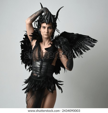 Close up portrait of female model wearing gothic horned headdress with halloween black dress and fantasy angel feather wings. Holding  bird prop with gestural arms posing. Isolated studio background