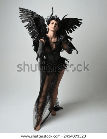 Full length portrait of female model wearing gothic horned headdress with halloween black dress and fantasy angel feather wings. Standing walking, holding pose bird prop. Isolated studio background