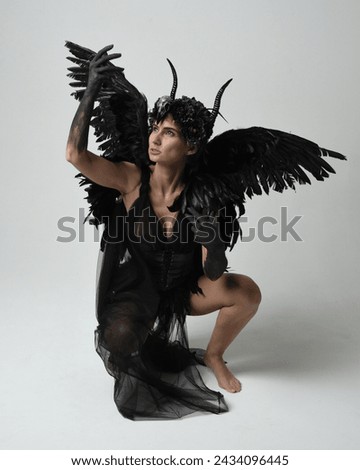 Full length portrait of female model wearing gothic horned headdress with halloween black dress and fantasy angel feather wings. Crouching pose, kneeling on floor. Isolated studio background