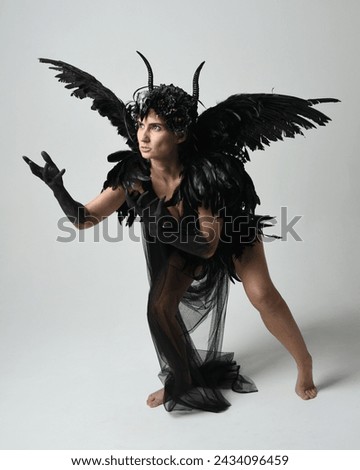 Full length portrait of female model wearing gothic horned headdress with halloween black dress and fantasy angel feather wings. Crouching pose, kneeling on floor. Isolated studio background
