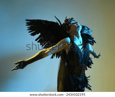 Close up portrait of female model wearing gothic horned headdress, halloween black dress and fantasy angel feather wings. Isolated dark studio background with cinematic shadow silhouettes 