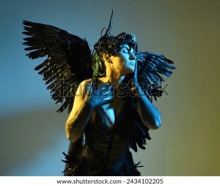 Close up portrait of female model wearing gothic horned headdress, halloween black dress and fantasy angel feather wings. Isolated dark studio background with cinematic shadow silhouettes 