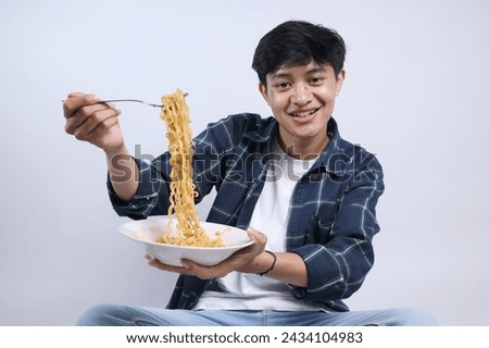 Young Asian man with plaid shirt, eating noodles with fork while sitting on the floor